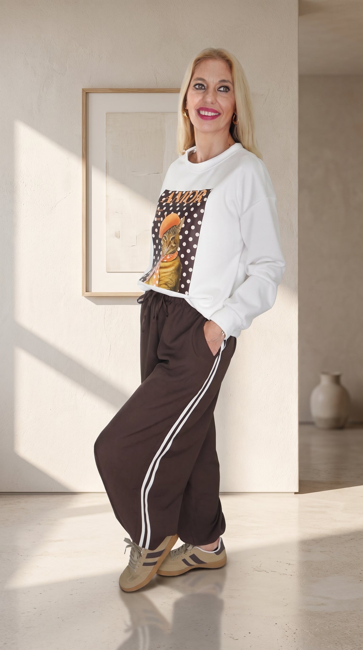 Vista lateral de modelo con sudadera blanca AMOR y pantalón Track marrón, con rayas laterales y bajos elásticos.